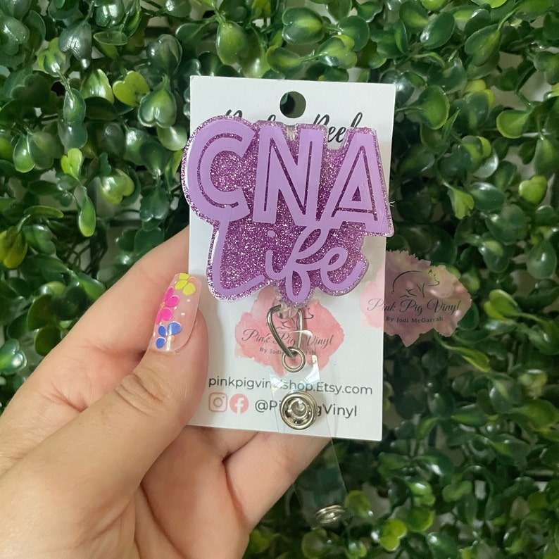 May include: A purple glitter badge reel with the words "CNA life" in a cursive font. The badge reel has a clear plastic retractable cord and a metal clip. The background is a green leafy plant.