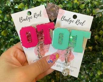 May include: Two pink and green glitter badge reels with the letters "OT" on them. The badge reels are attached to clear plastic retractable badge holders. The badge reels are on a white card with the text "Badge Reel" on it.