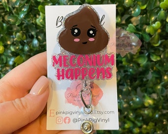 May include: A brown poop emoji with a pink glitter face and the words "MECONIUM HAPPENS" in pink glitter on a white background. The emoji is attached to a clear plastic badge holder with a silver clip.