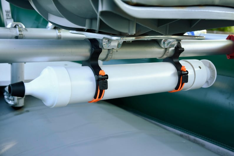 May include: A white cylindrical accessory with a black nozzle and a white cap, mounted on a boat. The accessory is secured with black and orange straps to a metal bar. The boat has a green and gray color scheme.