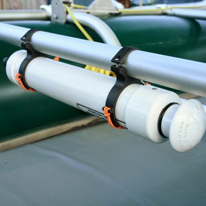 May include: A white cylindrical object with a rounded end is mounted on a silver metal bar with black and orange fasteners. The object is attached to a green surface, possibly a boat or raft.