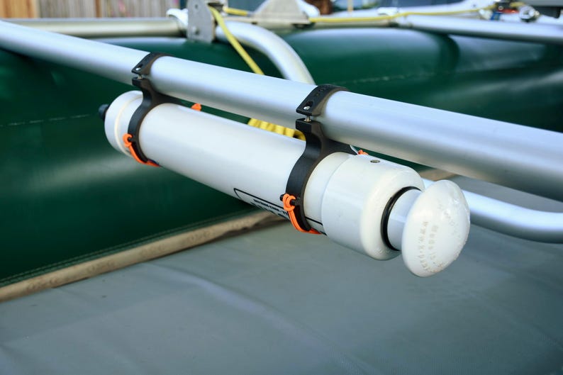 May include: A white cylindrical object with a rounded end is mounted on a silver metal bar with black and orange fasteners. The object is attached to a green surface, possibly a boat or raft.