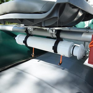 May include: A white cylindrical component is mounted on a boat, secured by black straps. The boat seat is gray, and a red container with an orange strap is visible. The boat's surface is green and gray.