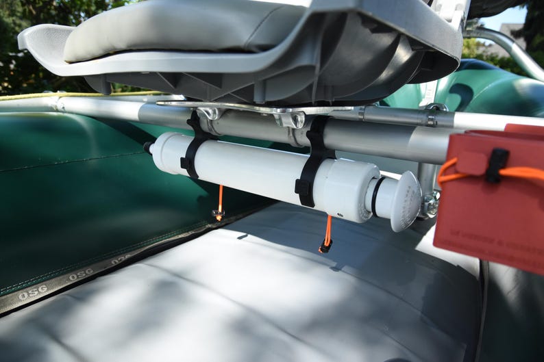 May include: A white cylindrical component is mounted on a boat, secured by black straps. The boat seat is gray, and a red container with an orange strap is visible. The boat's surface is green and gray.