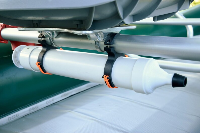 May include: A white cylindrical boat accessory with a black tip, secured by black and orange clamps to a silver bar. The accessory is mounted on a green and white boat structure.