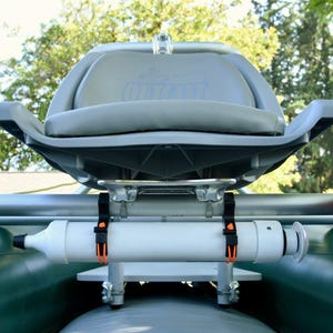 May include: A gray and white inflatable boat seat with a white cylindrical component and orange straps. The seat has a curved back and armrests. A red container with an orange cord is attached to the side.