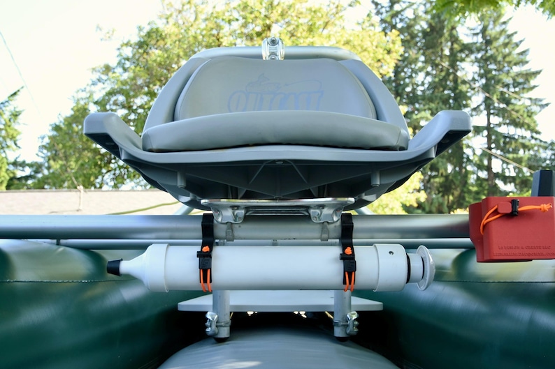 May include: A gray and white inflatable boat seat with a white cylindrical component and orange straps. The seat has a curved back and armrests. A red container with an orange cord is attached to the side.