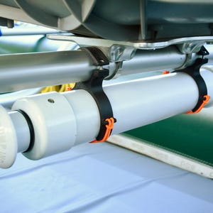 May include: A white cylindrical pump with a rounded handle, secured by black straps with orange accents to a silver bar. The pump is attached to a green surface, likely part of a boat or raft.
