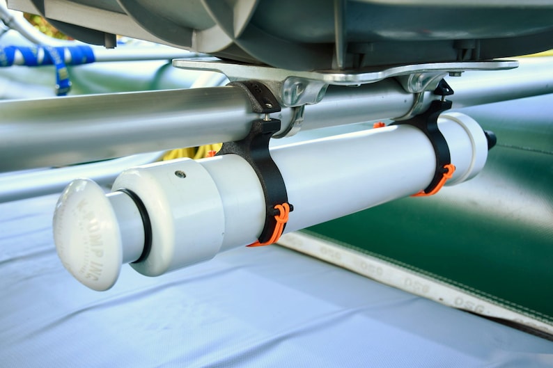 May include: A white cylindrical pump with a rounded handle, secured by black straps with orange accents to a silver bar. The pump is attached to a green surface, likely part of a boat or raft.