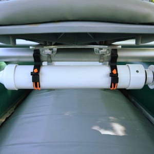 May include: A white cylindrical boat accessory is secured with black straps and orange buckles. The accessory is mounted below a gray boat seat and above a gray inflatable surface. The boat's green and red components are visible.