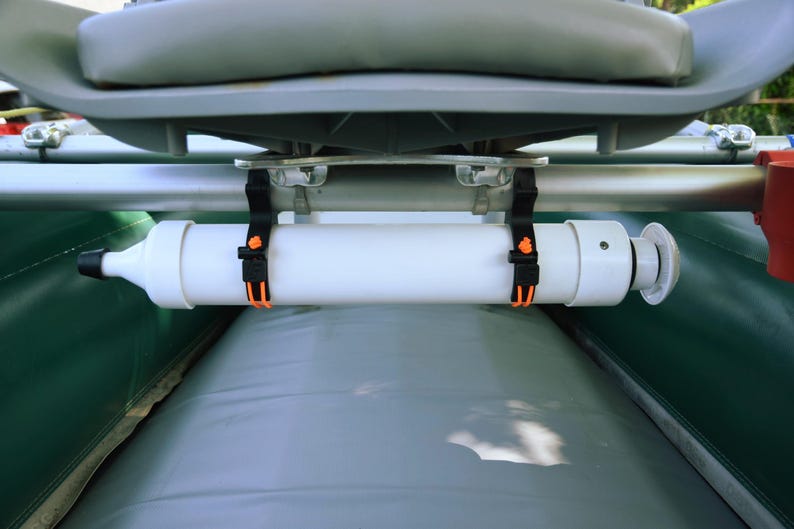 May include: A white cylindrical boat accessory is secured with black straps and orange buckles. The accessory is mounted below a gray boat seat and above a gray inflatable surface. The boat's green and red components are visible.