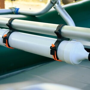 May include: A white cylindrical boat accessory with a black nozzle and orange straps, attached to a silver bar with black brackets. The accessory is mounted on a green surface, likely a boat.