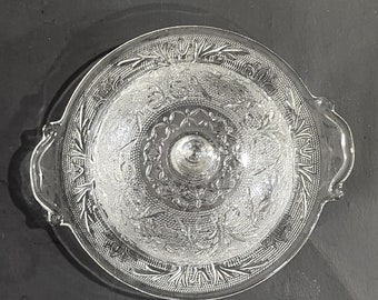 May include: A clear glass serving dish with a lid. The dish has a decorative etched pattern of flowers and leaves. The lid has two handles.