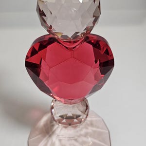 Op de afbeelding: Een decoratieve kristallen sculptuur met een gefacetteerd rood hart, een heldere bol en een heldere gefacetteerde top. De sculptuur staat op een heldere, gefacetteerde basis.