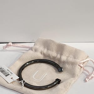 May include: A dark gray metal cuff bracelet with square ends rests on a light pink drawstring pouch. The bracelet is curved and has a minimalist design. A white tag with text is attached to the pouch.