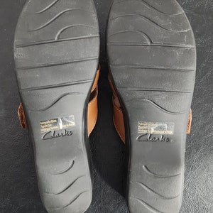 May include: A pair of tan leather sandals with black rubber outsoles featuring wavy tread patterns. The soles are marked with the Clarks brand name and a sticker stating Fabriqu&eacute; En Chine, Tige De Cuir, Mat&eacute;riaux Synth&eacute;tiques pour le Reste.