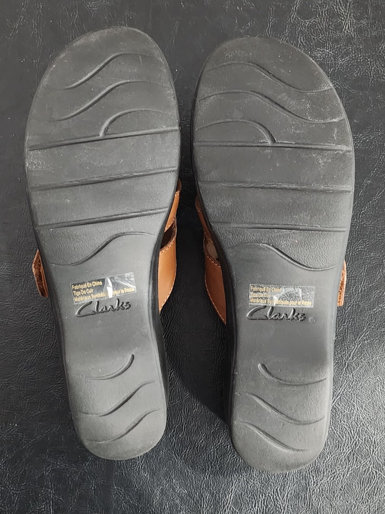 May include: A pair of tan leather sandals with black rubber outsoles featuring wavy tread patterns. The soles are marked with the Clarks brand name and a sticker stating Fabriqu&eacute; En Chine, Tige De Cuir, Mat&eacute;riaux Synth&eacute;tiques pour le Reste.