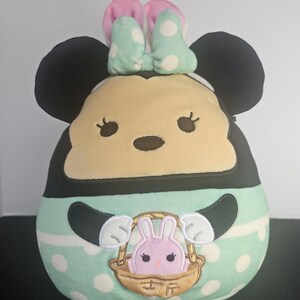 May include: A plush toy shaped like Minnie Mouse, featuring black ears, a tan face, and a mint green body with white polka dots. The toy has a pink and mint bow and a basket with a pink bunny.