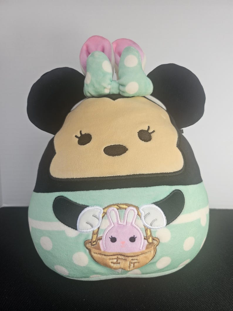 May include: A plush toy shaped like Minnie Mouse, featuring black ears, a tan face, and a mint green body with white polka dots. The toy has a pink and mint bow and a basket with a pink bunny.