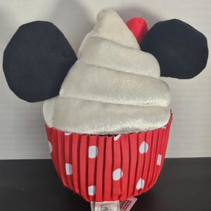 May include: A plush cupcake toy with black mouse ears and a red and white polka dot wrapper. The cupcake has a white frosting swirl and a red cherry on top. A tag is attached to the bottom.
