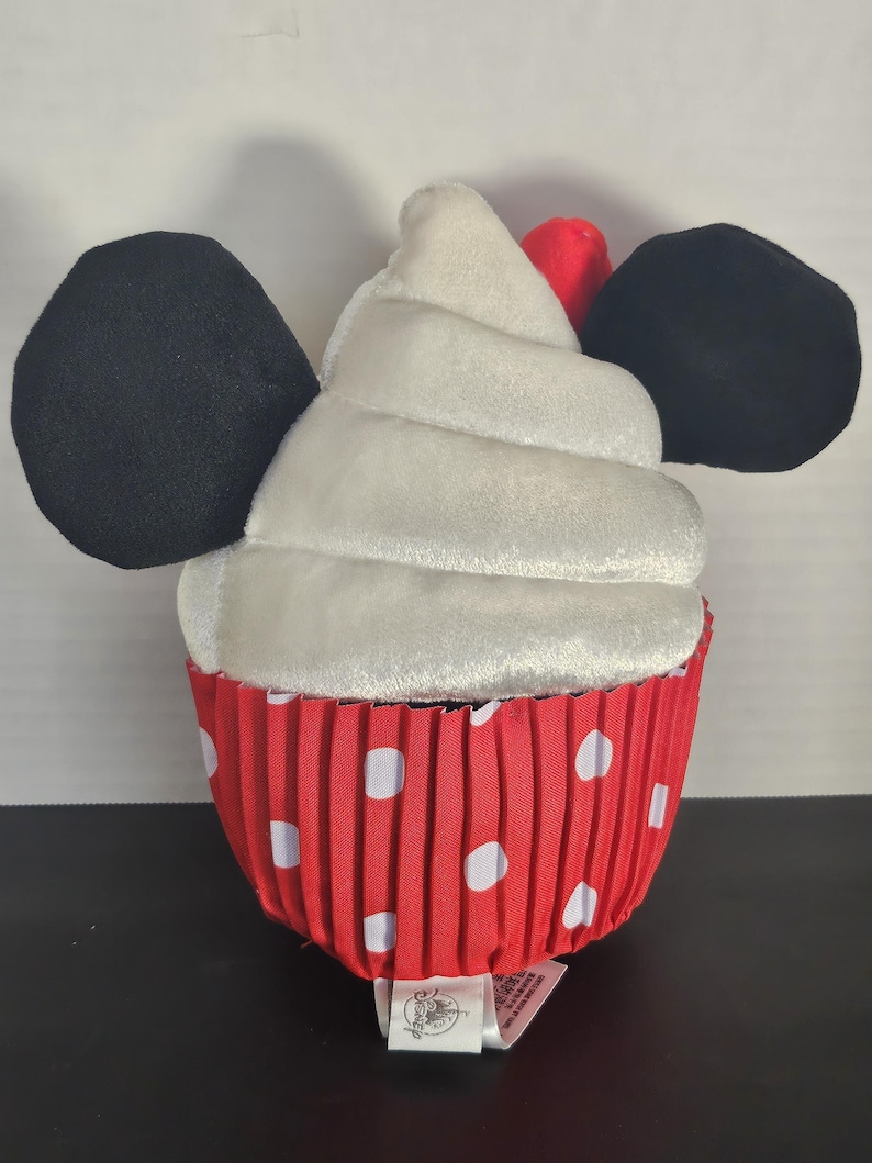 May include: A plush cupcake toy with black mouse ears and a red and white polka dot wrapper. The cupcake has a white frosting swirl and a red cherry on top. A tag is attached to the bottom.