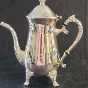 May include: A silver teapot with a decorative lid and handle. The teapot has three feet and a polished finish.