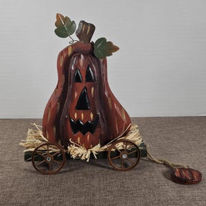 May include: A rustic, wooden pumpkin decoration with a carved face, mounted on a small wagon with straw. The pumpkin is painted in shades of brown and orange, with green leaves and a rope pull. A festive fall decoration.