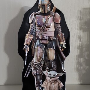 Puede incluir: Una fiambrera de metal negro con un personaje mandaloriano de la serie Star Wars. El Mandaloriano lleva un casco y una armadura y sostiene un arma. Un peque&ntilde;o personaje alien&iacute;gena verde est&aacute; de pie frente al Mandaloriano.