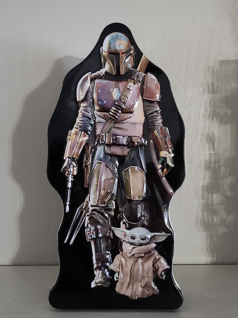 Puede incluir: Una fiambrera de metal negro con un personaje mandaloriano de la serie Star Wars. El Mandaloriano lleva un casco y una armadura y sostiene un arma. Un peque&ntilde;o personaje alien&iacute;gena verde est&aacute; de pie frente al Mandaloriano.