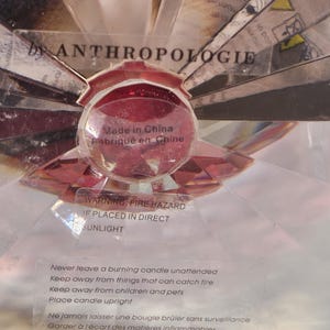 Op de afbeelding: Een decoratieve kandelaar met een rode, facetgeslepen glazen bol in het midden, omgeven door heldere, schuine glazen stukken. De tekst "by ANTHROPOLOGIE" is zichtbaar. Het label luidt "Made in China" en bevat een waarschuwing voor brandgevaar.