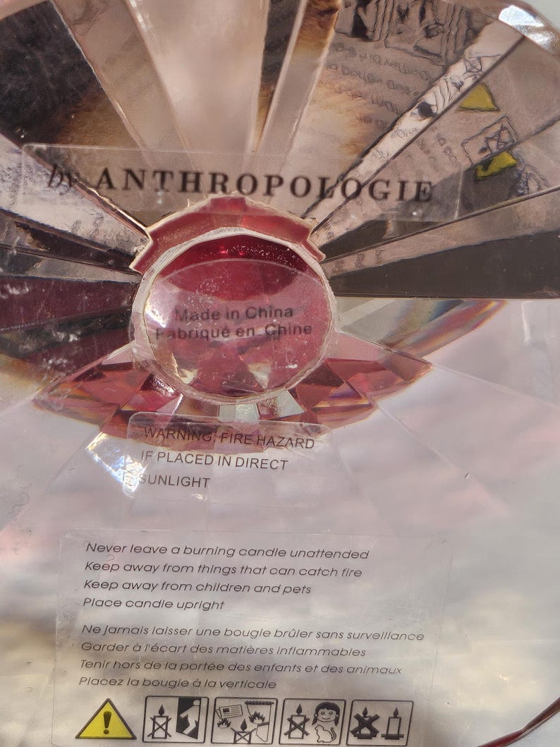 Op de afbeelding: Een decoratieve kandelaar met een rode, facetgeslepen glazen bol in het midden, omgeven door heldere, schuine glazen stukken. De tekst "by ANTHROPOLOGIE" is zichtbaar. Het label luidt "Made in China" en bevat een waarschuwing voor brandgevaar.