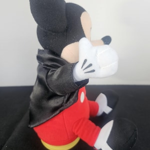 May include: A plush toy of a cartoon mouse character wearing a black satin tuxedo jacket, red trousers with white buttons, and white gloves. The character has black ears, a tan face, and black shoes with white accents, shown in a seated position.