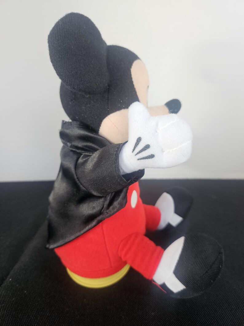 May include: A plush toy of a cartoon mouse character wearing a black satin tuxedo jacket, red trousers with white buttons, and white gloves. The character has black ears, a tan face, and black shoes with white accents, shown in a seated position.