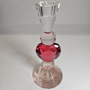 Op de afbeelding: Een heldere kristallen kandelaar met een facetontwerp. De kandelaar heeft een rood hartvormig kristal in het midden, met heldere kristallen bollen erboven en eronder. De basis is een lichtroze, gefacetteerde cirkel.