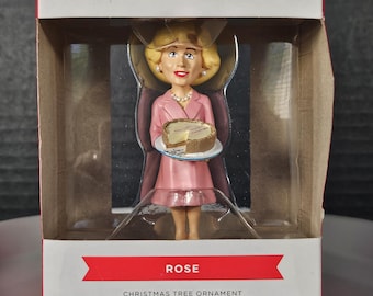 Könnte beinhalten: Ein Hallmark-Anhänger mit Rose aus der Fernsehserie Golden Girls. Rose hält einen Teller mit einem Stück Käsekuchen. Der Anhänger ist in einer rot-weißen Schachtel verpackt, auf der "The Golden Girls" und "Hallmark Ornaments" steht.