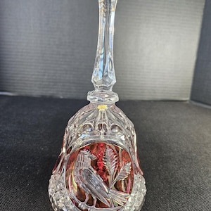 May include: A clear crystal bell with a long, faceted handle. The bell features a detailed carving of a bird and foliage, set against a red background. The bell has a diamond-cut pattern and a decorative top.