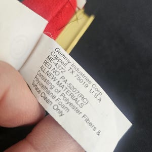 May include: A white fabric product tag with black text reading Gemmy Industries Corp. Coppell, TX 75019 U.S.A ME-4372 REG NO PA-5207(RC) ALL NEW MATERIALS Consisting of Polyester Fibers & Polyurethane Foam Surface Clean Only, attached to a red object.