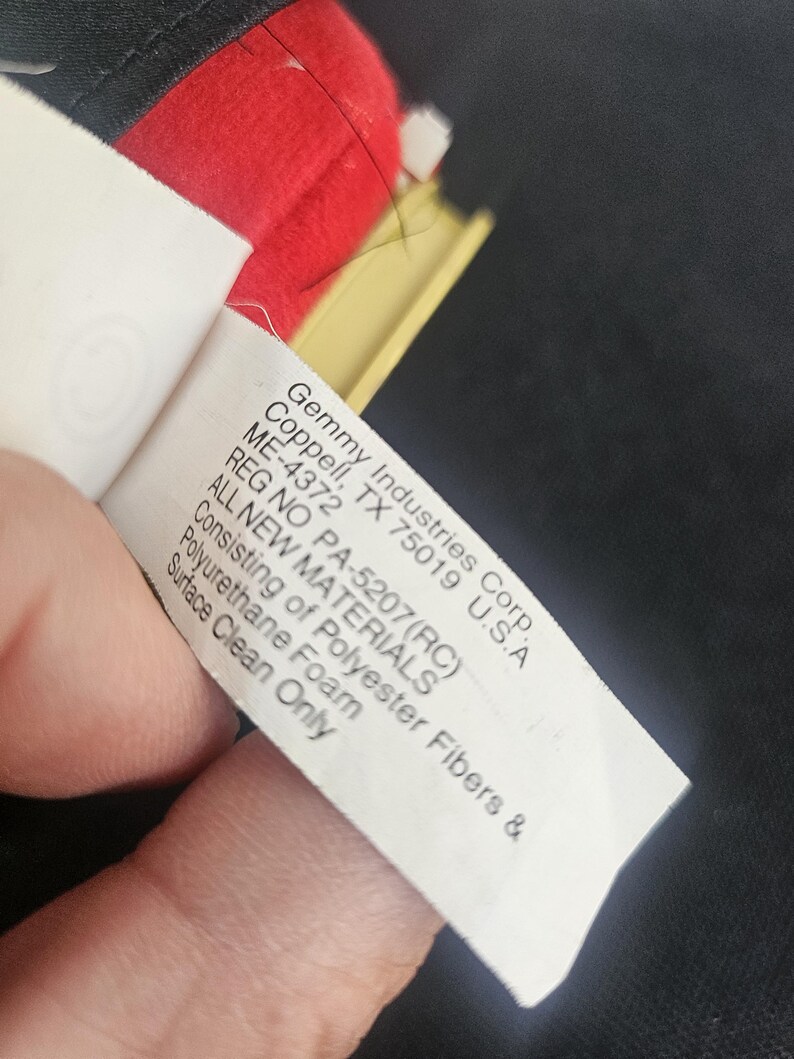 May include: A white fabric product tag with black text reading Gemmy Industries Corp. Coppell, TX 75019 U.S.A ME-4372 REG NO PA-5207(RC) ALL NEW MATERIALS Consisting of Polyester Fibers & Polyurethane Foam Surface Clean Only, attached to a red object.