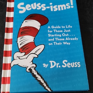 May include: A blue book with a red and white striped spine. The cover features a cartoon cat wearing a red and white hat with a red bow. The title of the book is "Seuss-isms!" and the subtitle is "A Guide to Life for Those Just Starting Out... and Those Already on Their Way." The author is "By Dr. Seuss."