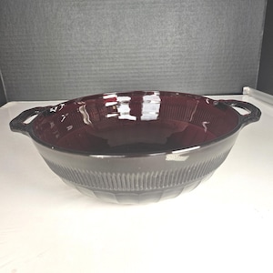 May include: A dark burgundy glass bowl with two handles. The bowl has a ribbed design around the exterior and a smooth interior. The bowl is sitting on a white surface.