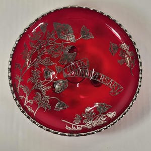 May include: A round, red glass dish with a silver rim and floral design. The dish has the words "40th Anniversary" on a banner. The floral design is silver and covers the surface of the dish.