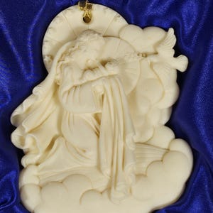 May include: Ivory-colored angel figurine with a halo, dove, and flowing robes. The angel is seated on a cloud, with a gold-colored accent at the top. The figurine is set against a blue background.