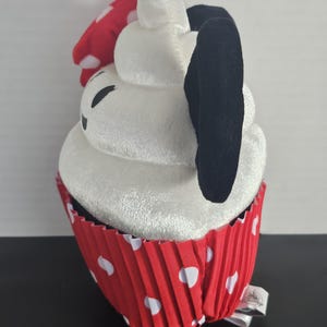 May include: A plush cupcake toy with a white frosting top, black ears, and a red polka-dot bow. The cupcake sits in a red paper liner with white polka dots. A small tag is attached to the bottom.