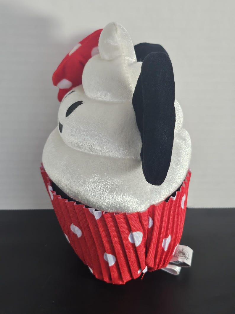 May include: A plush cupcake toy with a white frosting top, black ears, and a red polka-dot bow. The cupcake sits in a red paper liner with white polka dots. A small tag is attached to the bottom.
