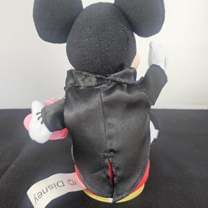 May include: A plush Mickey Mouse figure viewed from the back, wearing a black satin tuxedo jacket with a tail. The figure has large round black ears, white gloves, and a white fabric tag extending from the base that reads Disney in black text.