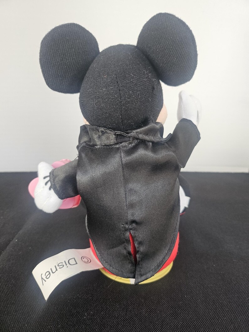 May include: A plush Mickey Mouse figure viewed from the back, wearing a black satin tuxedo jacket with a tail. The figure has large round black ears, white gloves, and a white fabric tag extending from the base that reads Disney in black text.