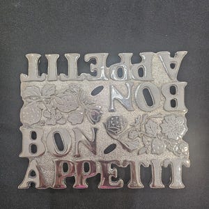 May include: A rectangular silver-toned metal trivet featuring the repeated text BON APPETIT in raised, polished lettering. The design includes embossed strawberry and floral motifs with a textured, matte background finish between the letters.