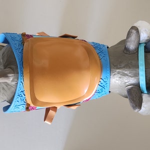 Pu&ograve; includere: Una renna giocattolo grigia con un'imbracatura azzurra e una sella marrone. La renna ha orecchie bianche e soffici e una cinghia azzurra attorno alle corna. L'imbracatura ha un motivo blu decorativo.