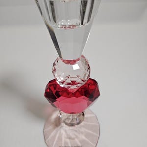 Op de afbeelding: Een heldere kristallen kandelaar met een facetontwerp. De kandelaar heeft een rood hartvormig kristal in het midden, met een rond kristal erboven en een uitlopende top. De basis is een helder, veelzijdig ontwerp.