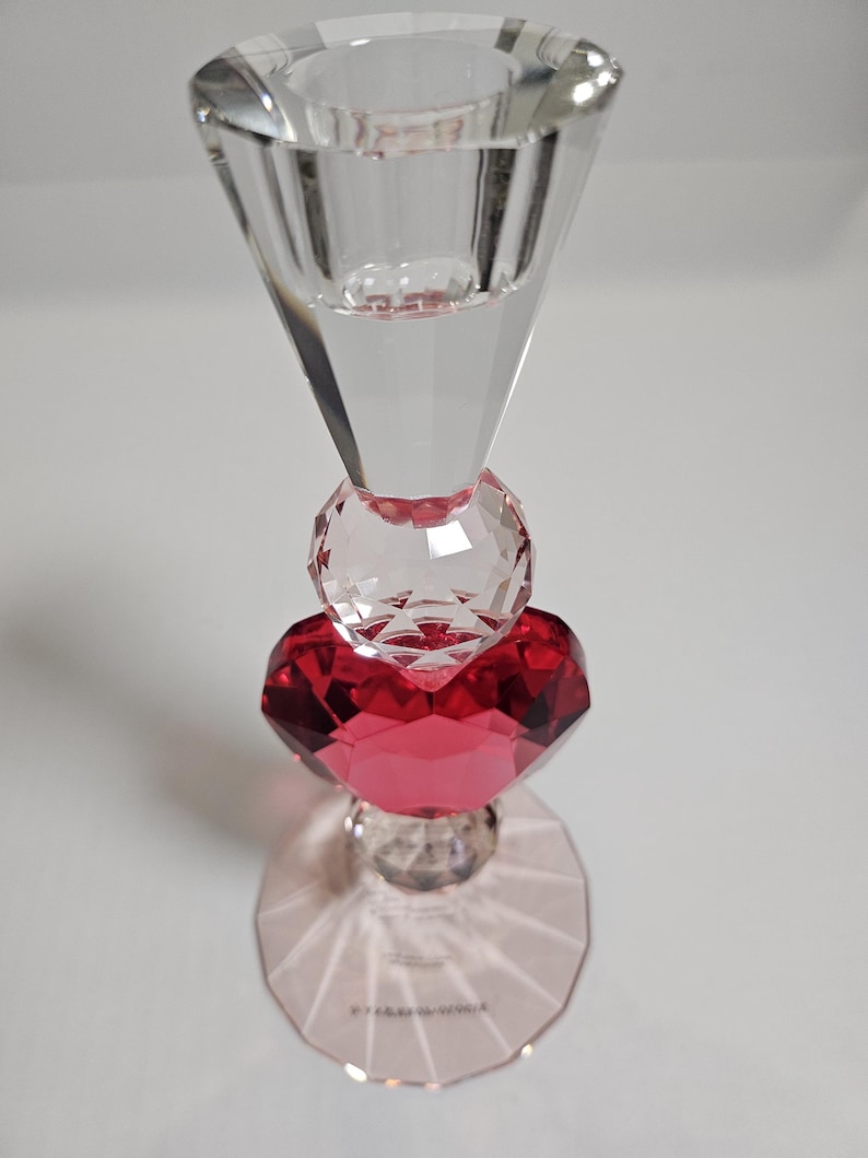 Op de afbeelding: Een heldere kristallen kandelaar met een facetontwerp. De kandelaar heeft een rood hartvormig kristal in het midden, met een rond kristal erboven en een uitlopende top. De basis is een helder, veelzijdig ontwerp.